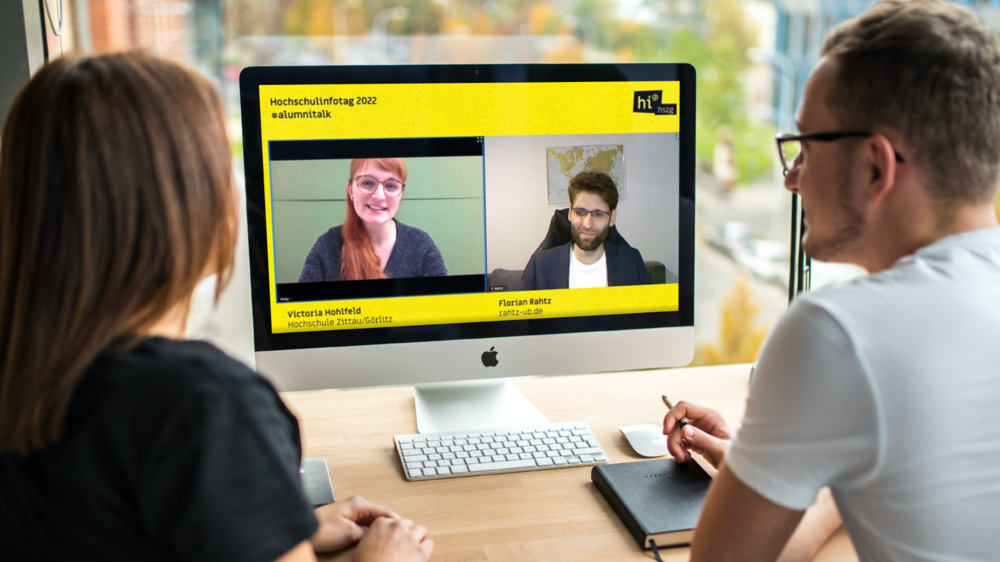 Zwei Studierende sitzen vor einem PC. Auf dem Bildschirm ist eine Moderatorin und ein Gast im Gespräch zu sehen.