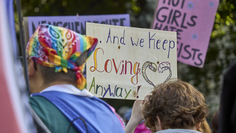 Ein CSD-Teilnehmer mit Regenbogenfarbigen Kopftuch und eine Begleitung laufen beim Christopher Street Day mit. Man sieht ihre Rückenansicht. Dazwischen ist ein Schild mit der Aufschrift: And we keep loving anyway, zu lesen.