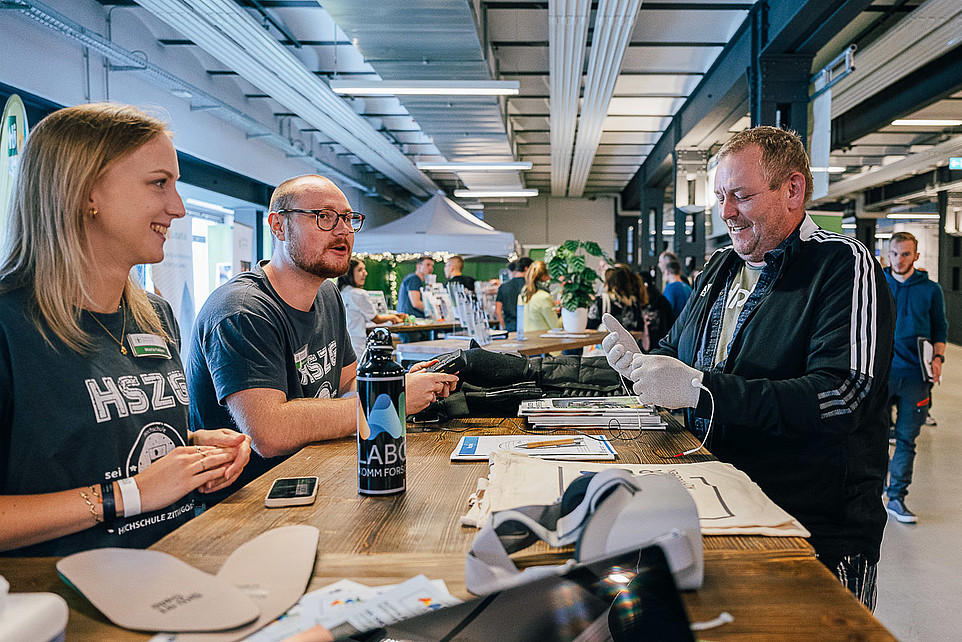 Drei Leute unterhalten sich wobei einer Tremorhandschuhe austestet