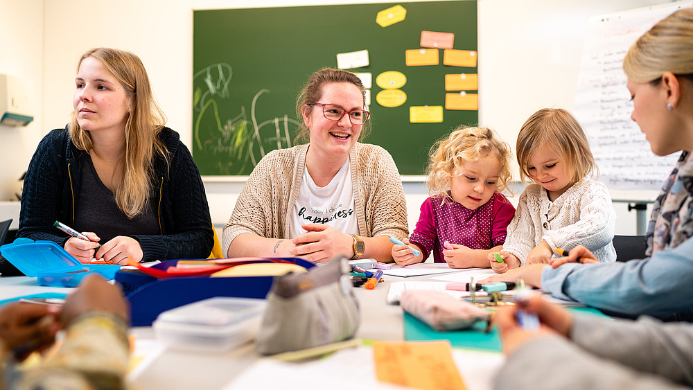 Lehrende und Kinder am Tisch, im Hintergrund eine grüne Tafel mit Zetteln.