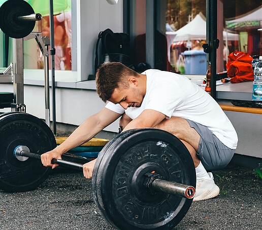 Ein Student kniet vor einer Langhantel und umschließt mit seinen Händen die Hantelstange.