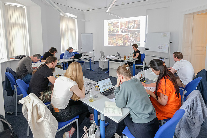 In einem Seminarraum sitzen Kursteilnehmende und hören der Seminarleiterin zu, die etwas anhand einer Beamerprojektion erklärt.