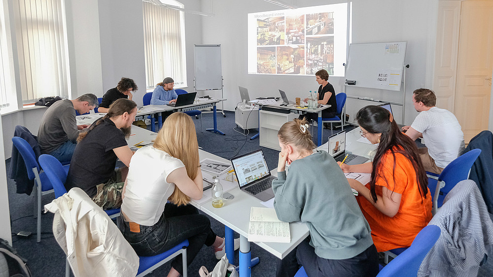 In einem Seminarraum sitzt eine Gruppe von Kursteilnehmenden und macht sich Notizen. Am Tisch sitzt die Dozentin und erklärt die Kursinhalte anhand der Beamerprojektion.