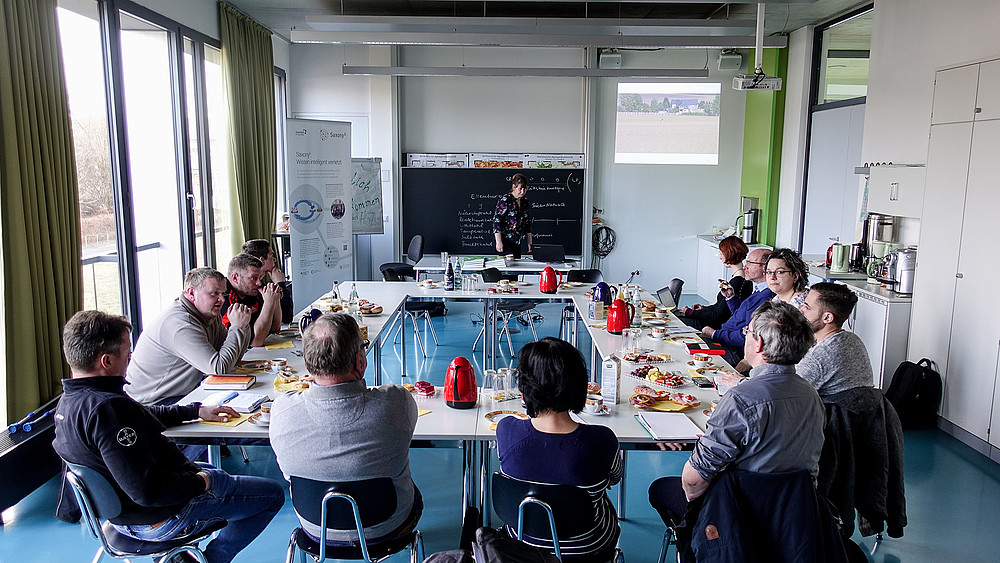 Die Teilnehmer*innen sitzen zusammen an einem Tisch vor einer Tafel und unterhalten sich.an einem 