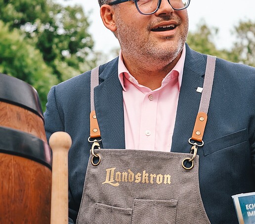 Der Rektor trägt eine Bierschürze und steht neben dem Bierfass.