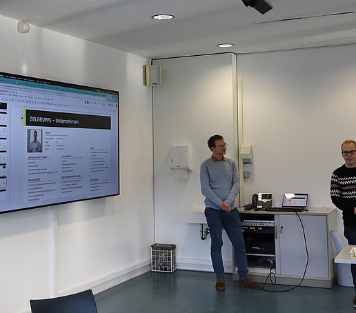 Workshopteilnehmende stehen im Seminarraum vor einem großen Monitor.