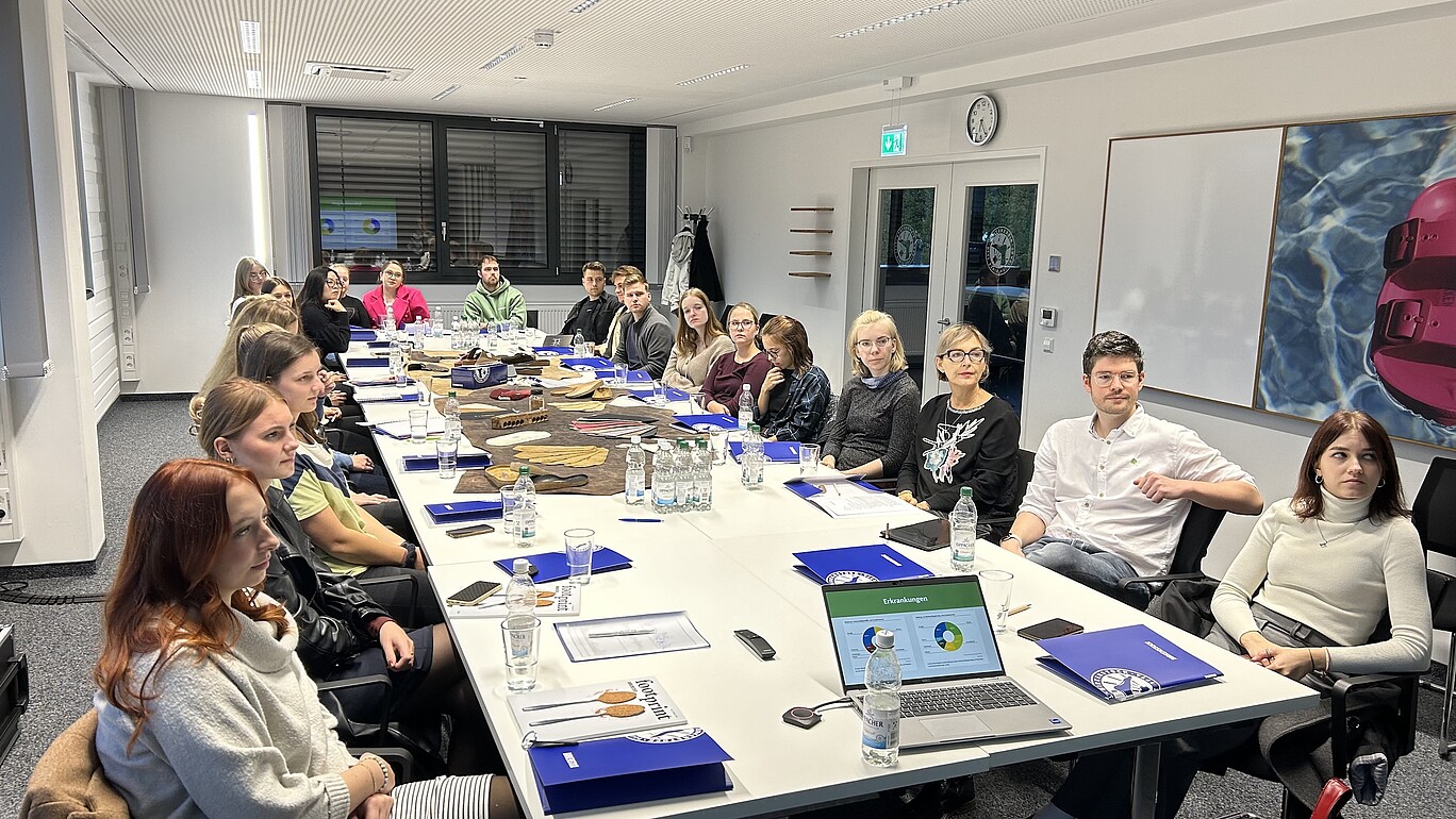 Das Bild zeigt eine Gruppe von etwa 18 jungen Erwachsenen, die um einen langen Konferenztisch in einem modernen Büro sitzen und an einer Besprechung teilnehmen.   Das Bild zeigt eine Gruppe von etwa 18 jungen Erwachsenen, die um einen langen, weißen Konferenztisch in einem hellen, modernen Büro sitzen. Die Teilnehmer scheinen an einer Besprechung oder einem Workshop teilzunehmen. Sie sind in informeller Kleidung gekleidet, einige tragen Jacken, andere Sweatshirts oder Hemden.  Auf dem Tisch befinden sich zahlreiche Gegenstände: Wasserflaschen, Gläser, Notizbücher, Stifte, Präsentationen auf Laptops, sowie einige Materialen, die auf eine Art von Workshop hindeuten (z.B. Lederproben). Einige Teilnehmer schauen auf den Laptop, andere untereinander oder in den Raum. Die Atmosphäre wirkt konzentriert und professionell, aber auch entspannt und freundlich. Die Beleuchtung ist hell und künstlich, die Wände sind weiß gestrichen. Ein großes Kunstwerk mit einem pinken Gegenstand im Wasser ist an der Wand zu sehen.  Das Gesamtbild wirkt modern, hell und freundlich. Es vermittelt den Eindruck einer dynamischen und professionellen Arbeitsumgebung.