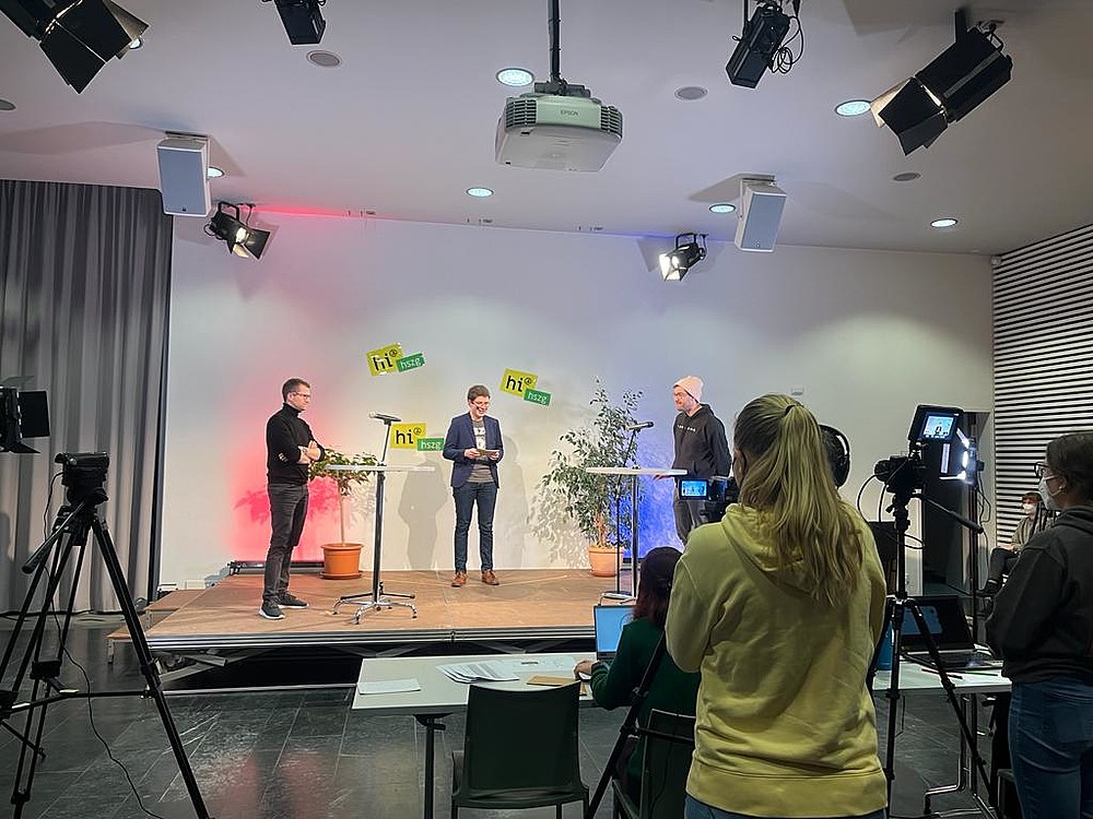 Blick auf eine Bühne mit drei Personen. dazu Scheinwerfer und Kameras die auf die Bühne gerichtet sind.