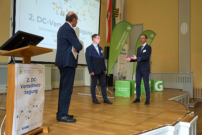 Drei Männer im Anzug diskutieren auf der Bühne vor Veranstaltungsbanner.