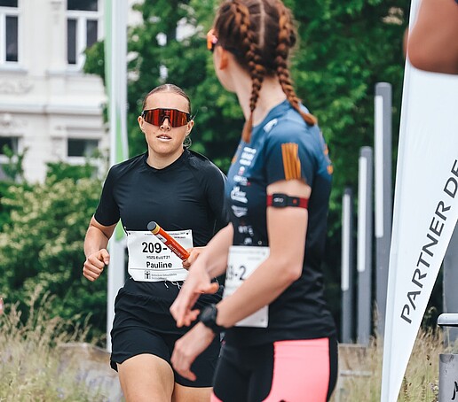 Eine Läuferin läuft mit Staffelstab auf eine wartende Läuferin zu.