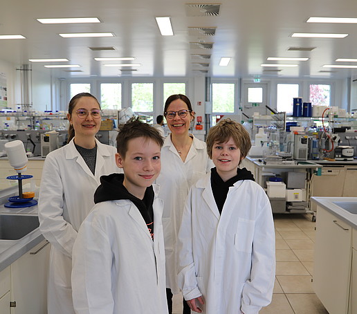 Eine Labormitarbeiterin, eine Professorin und zwei Schüler in weißen Kitteln stehen im Labor für pharmazeutische Biotechnologie und lächeln in die Kamera.