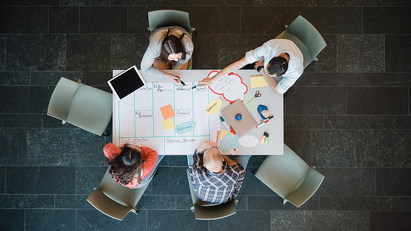 An einem Tisch sitzen vier Studierende. Unterschiedliche Arbeitsmaterialien wie ein Plakat, Notizzettel und ein Tablet liegen vor ihnen auf dem Tisch. Sie unterhalten sich.