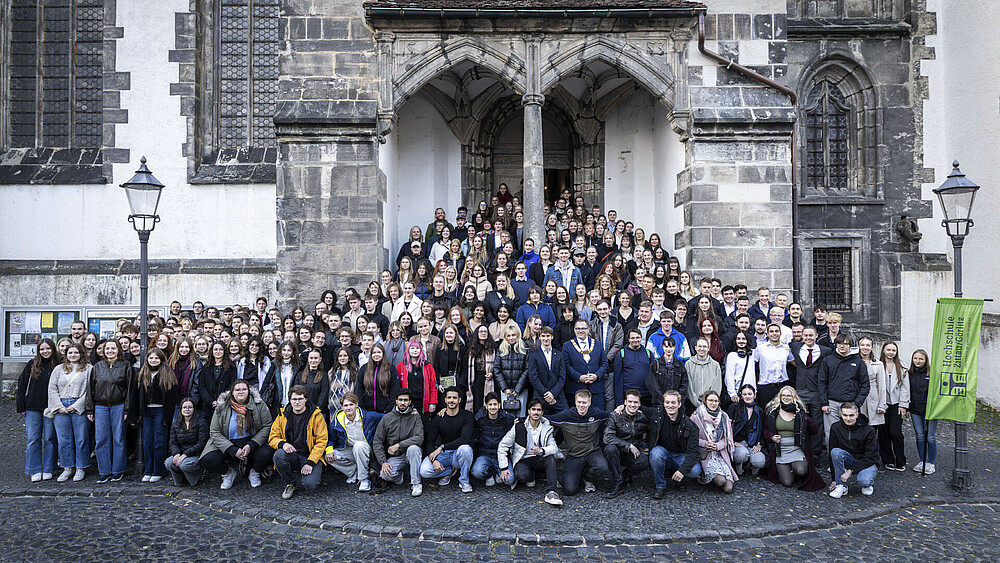 Eine große Menge junge Studierende und der Rektor stehen am Seiteneingang der Kirche auf den Stufen und dem vorderen Platz verteilt. Alle schauen in die Kamera.