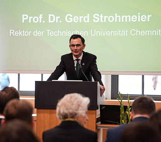Ein Mann im dunklen Anzug steht an einem Rednerpult und spricht zu einem Publikum. Im Hintergrund ist eine Leinwand mit der Aufschrift „Prof. Dr. Gerd Strohmeier, Rektor der Technischen Universität Chemnitz“ zu sehen.