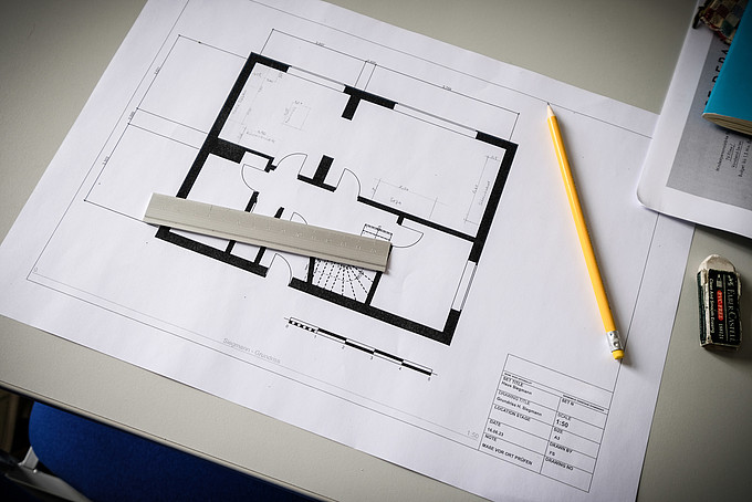 Auf einem Seminartisch liegt ein Grundriss von einer Wohnung. Darauf liegt ein gelber Bleistift und ein Lineal.