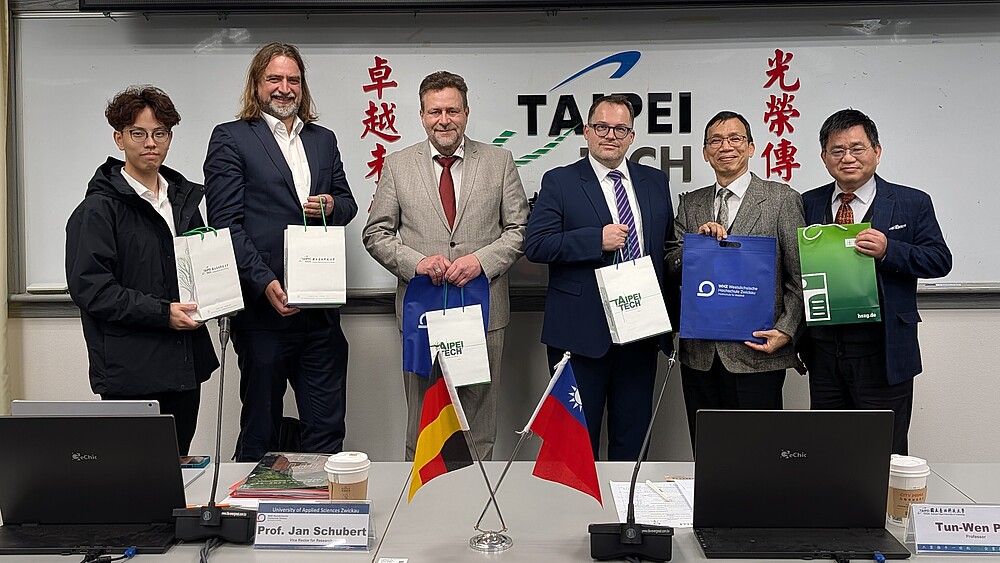 Rektor Alexander Kratzsch und Mitglieder der sächsischen Delegation stehen gemeinsam mit Vertretern der Taipei Tech Universität vor einem Whiteboard und halten Gastgeschenke in den Händen. Auf dem Tisch stehen deutsche und taiwanesische Flaggen.