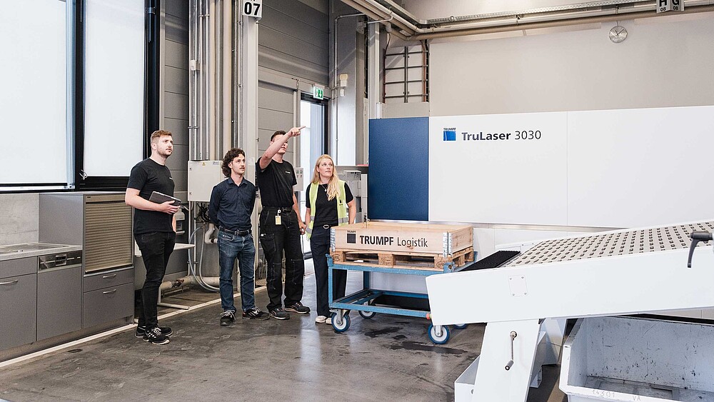 In einer Maschinenhalle stehen drei Männer und eine Frau. Ein Mann zeigt mit ausgestrecktem Arm auf eine Industrieanlage.