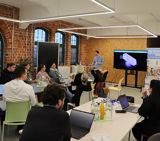 Workshopteilnehmende sitzen am Tisch und hören einem Vortragenden zu. 