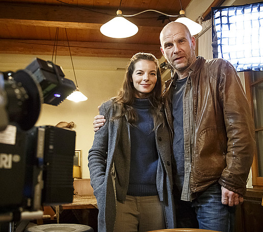 Eine Schauspielerin und ein Schauspieler posieren auf einem Filmset vor einer Kamera.