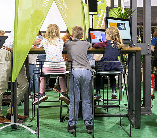 Drei junge Schüler sitzen und stehen an einem Hochtisch. Man sieht sie von hinten.