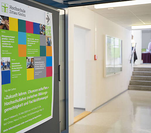 Ein Poster hängt an einem grauen Ständer, daneben ein Durchgang im Gebäude.