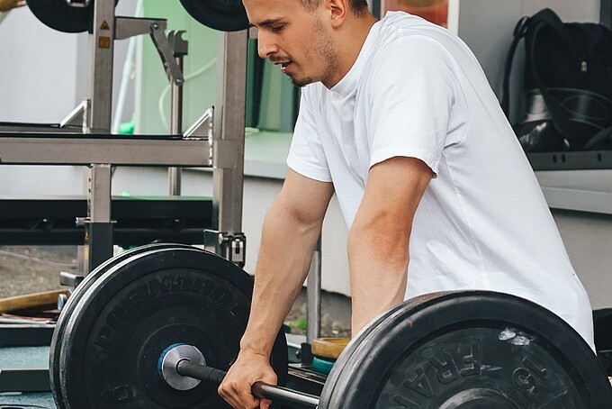 Ein junger Mann kniet und ist dabei eine schwere Langhantel zu stemmen.