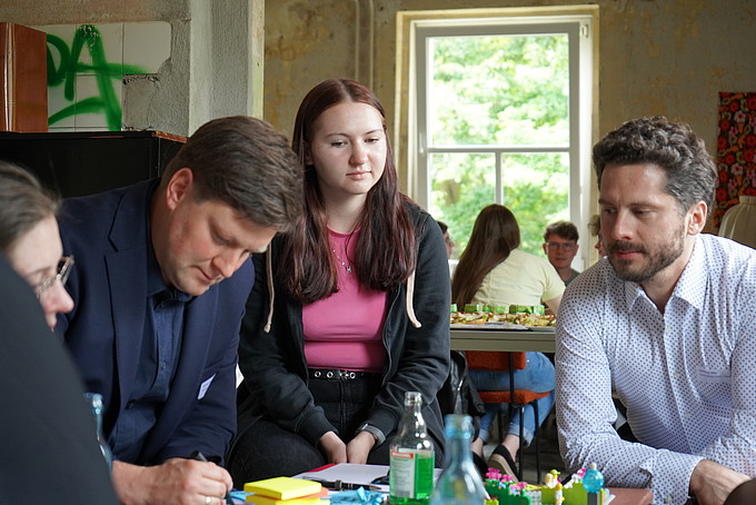 Eine Gruppe von Teilnehmenden sitzt an einem Tisch und diskutiert, während ein Moderator Notizen macht