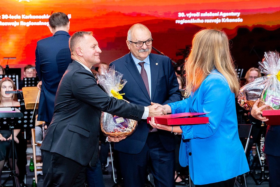 Teilnehmende auf der Bühne halten einen Präsentkorb in der Hand und schütteln einer Dame mit blauem Blazer die Hand.