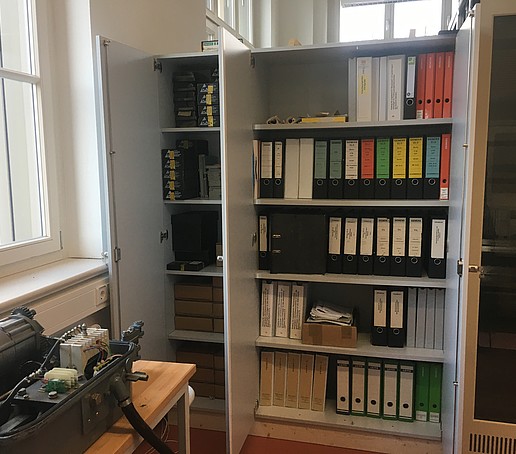 Ein Regal mit vielen Aktenordnern sowie ein Tisch mit verschiedenem Material