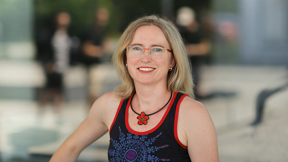 Dr. rer. nat. Kateřina Barková, eine blonde Frau mit Brille im sommerlichen Top mit roter Blumenhalskette, lächelt in die Kamera.