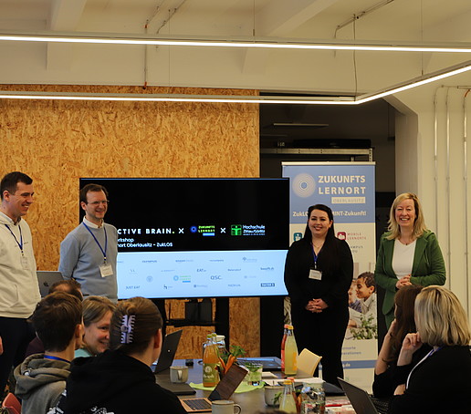 Workshopteilnehmende sitzen im Celsiuz am Tisch und vier Personen stehen vor einem Bildschirm und erläutern den nächsten Tagesordnungspunkt.