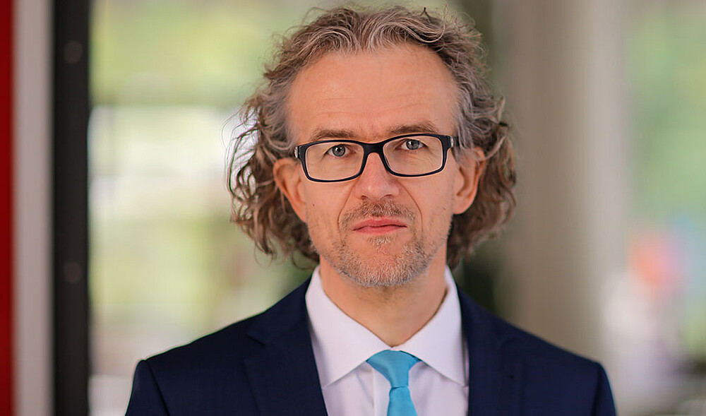 Prof. Saatkamp trägt Brille und eine hellblaue Krawatte und schaut in die Kamera.