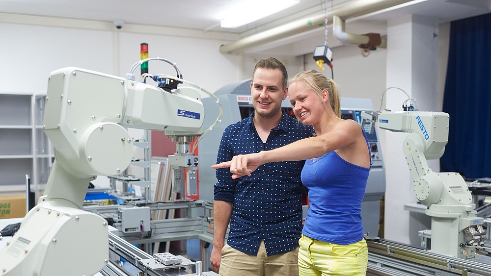 Zwei junge KIA-Studierende stehen im Robotik-Labor und schauen sich die Technik an.