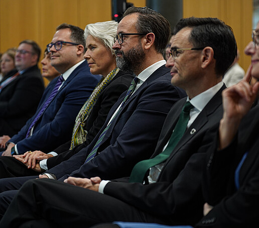 Mehrere Personen sitzen in einer Reihe und hören aufmerksam einer Veranstaltung zu.