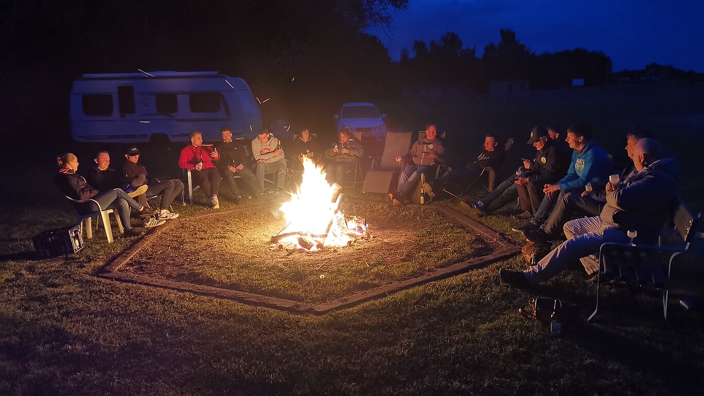 Studierende sitzen rund um ein Lagerfeuer auf einem Feld.