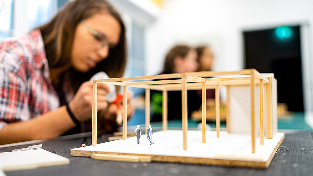 Eine Studentin benutzt Bastelkleber, um ein Hausmodell aus Holzstäben zu bauen. Das Modell steht vor ihr auf dem Tisch.