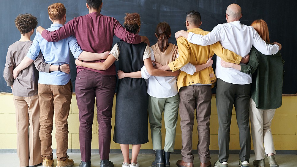 Eine Reihe von junger Menschen unterschiedlicher ethnischer Herkunft stehen in einer Reihe, den Rücken zur Kamera gedreht, mit Blick auf eine Tafel. Sie legen sich gegenseitig die Hände und Arme auf den jeweiligen Danebenstehenden.