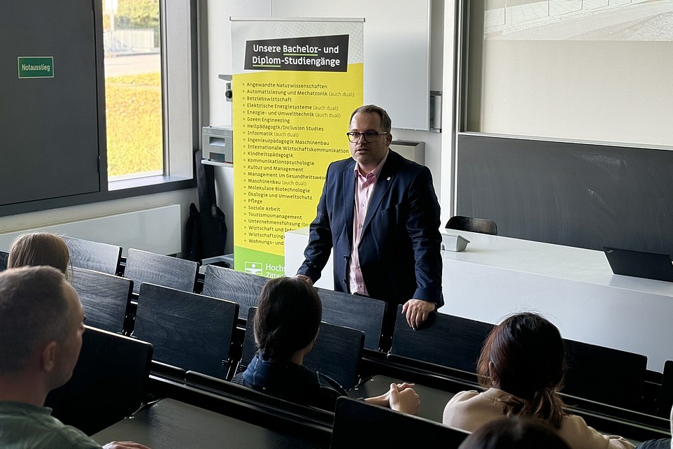 Der Rektor der HSZG, Prof. Dr.-Ing. Alexander Kratzsch, steht vor einer Gruppe von Studieninteressierten des Rahn Studienkollegs Leipzig und begrüßt sie im Hörsaal. Im Hintergrund sind ein Banner mit Studienangeboten und eine Präsentationsleinwand zu sehen.