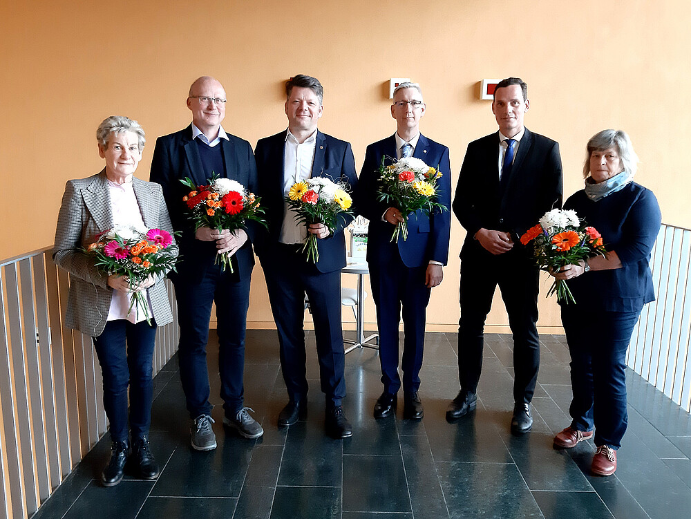 Sechs Personen des Hochschulrates stehen im Flur, halten Blumensträuße und lächeln in die Kamera.