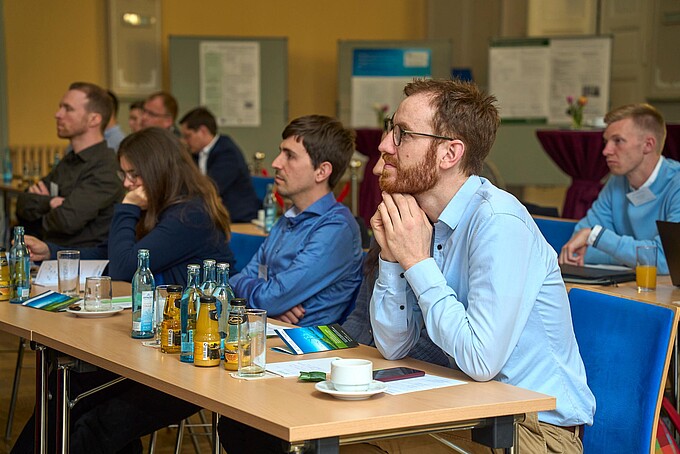 Teilnehmende sitzen aufmerksam an Tischen mit Getränken und Tagungsunterlagen.