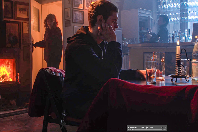 In einer Kulisse sitzt ein junger Mann an einem Tisch mit einem leeren Glas vor sich stehend.