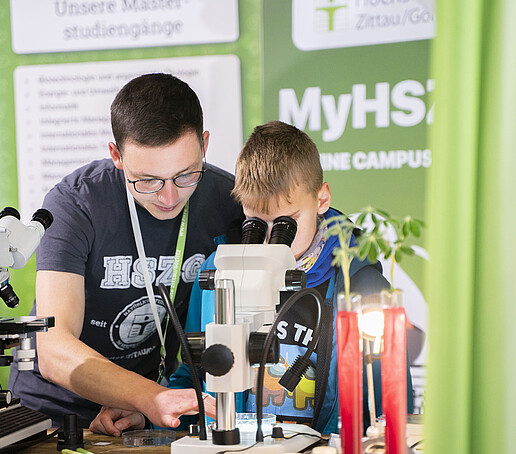 Ein Junge schaut durch ein Mikroskop, eine Studentische Hilfskraft leitet ihn an.