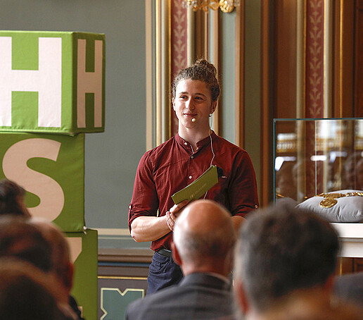 Der Moderator spricht dem Publikum zugewandt. In einem Glaskasten rechts daneben liegt die goldene Amtskette.