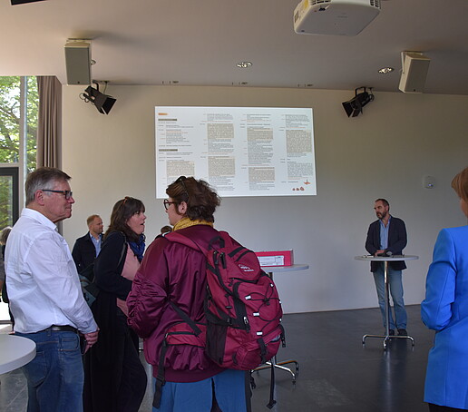 Teilnehmer in der Aula schauen auf die Projektionswand und hören einem Redner zu
