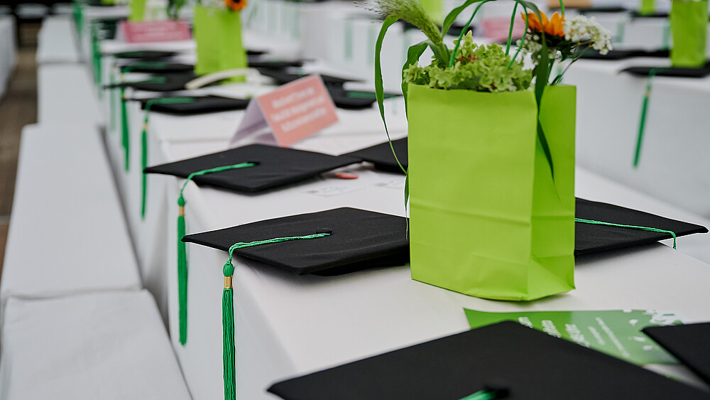 Auf einem mit weißer Tischdecke bedeckten Tisch liegen Absolventenhüte und in grüne Papiertüten gesteckte Blumensträuße.