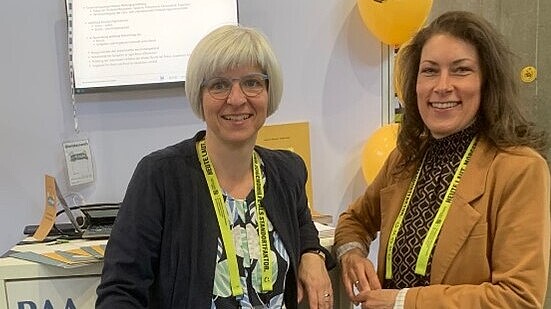 Zwei Frauen stehen vor einem Monitor und lächeln in die Kamera.