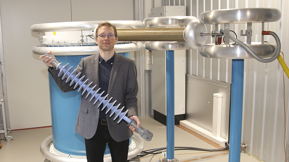 Ein Professor steht vor einer Spannungsanlage und hält ein Bauteil in der Hand.