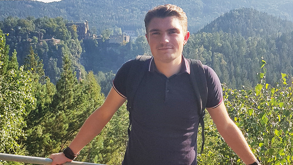 Franz Wenger steht auf einer Aussichtsplattform im Gebirge. Im Hintergrund sieht man eine Burg und eine Berglandschaft.