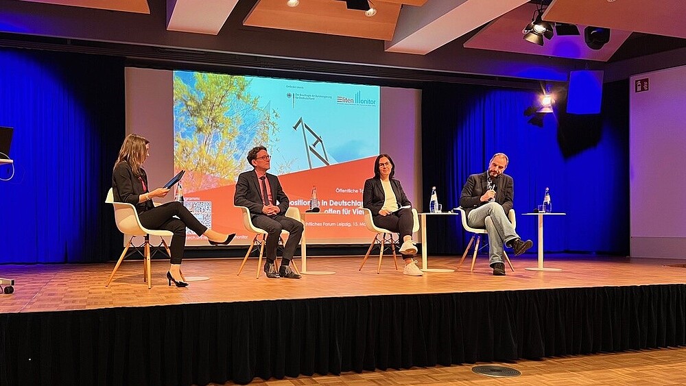 Vier Menschen sitzen auf einem Podium und diskutieren. Im Hintergrund leuchtet eine Projektion.