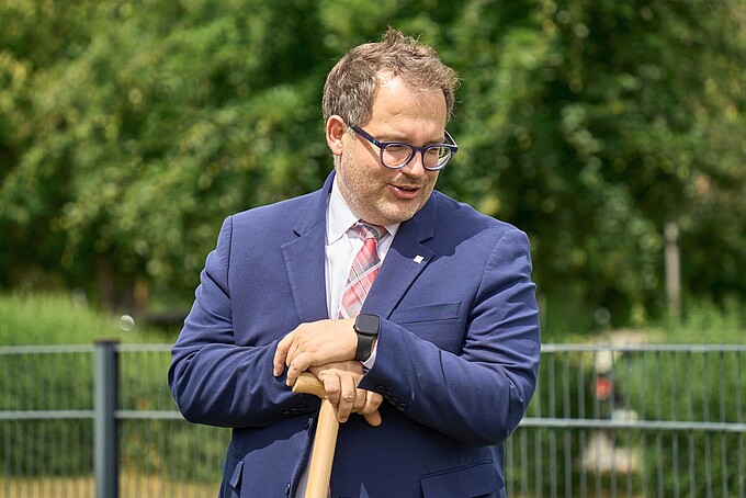 Der Rektor stützt sich auf eine Schaufel und schaut zu Boden.